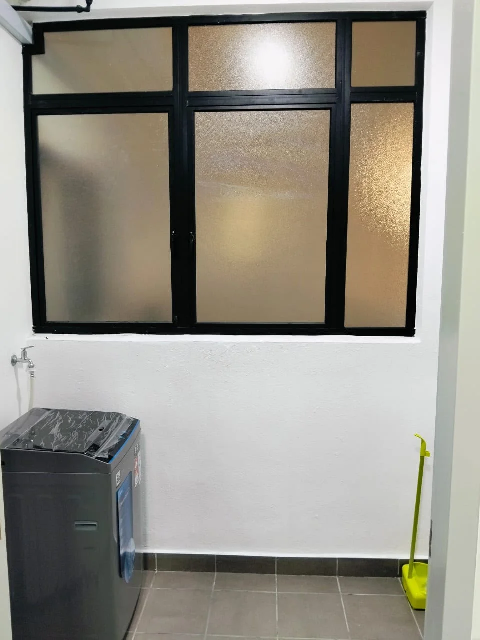 Laundry features: washing machine, black framed window, white painted walls, grey tiled flooring, floor drain, yellow mop, sink connection, natural light from window.